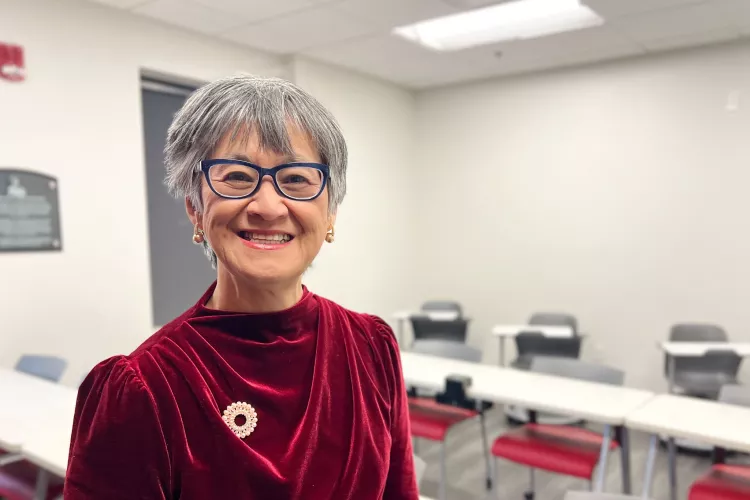 UofL School of Nursing Professor Dr. Ratchneewan Ross smiling at the camera in a classrrom. Student rows fo desks and chairs are behind her.